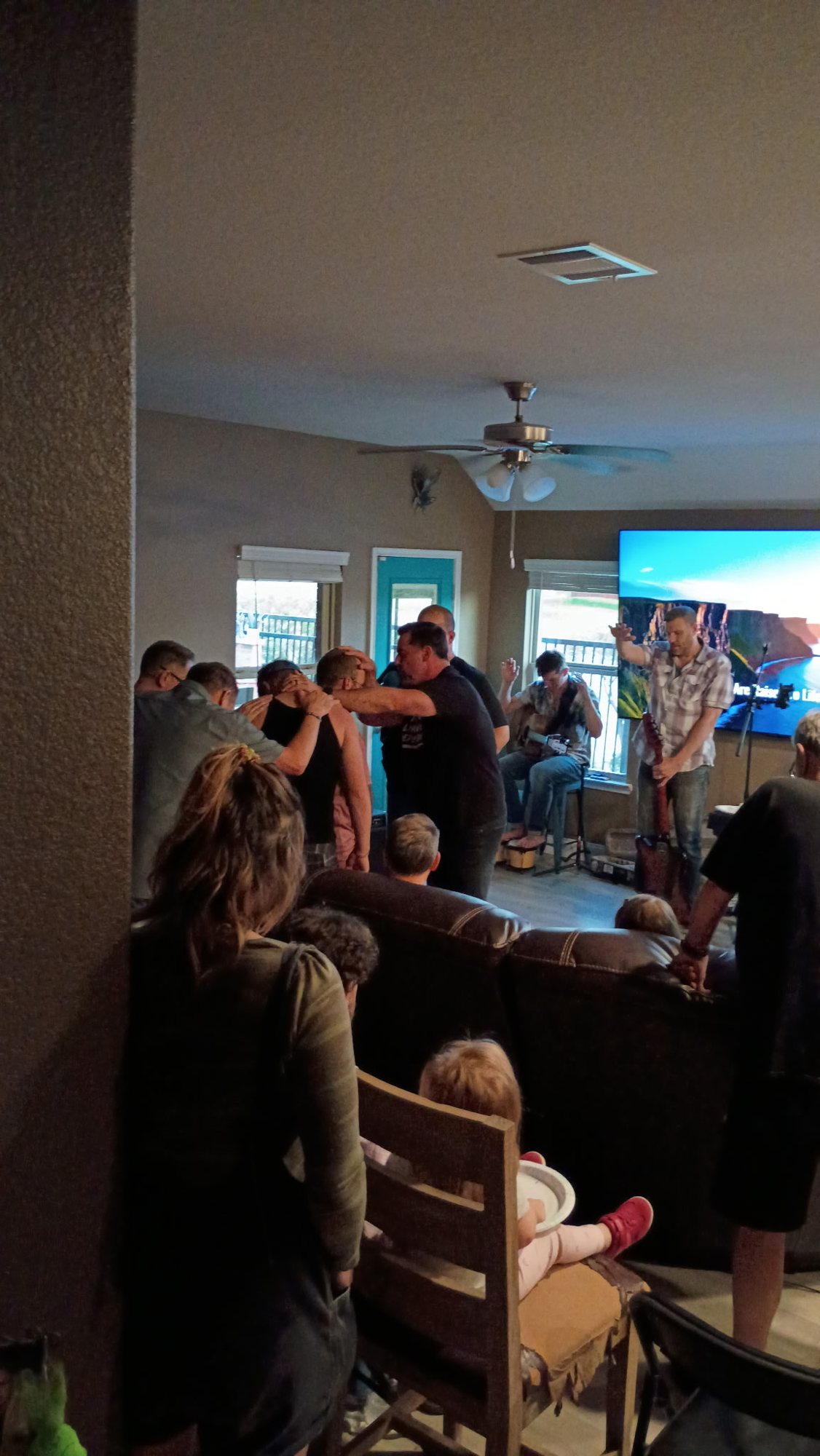 A worship night in a living room — hands laid on a brother in prayer while the band plays